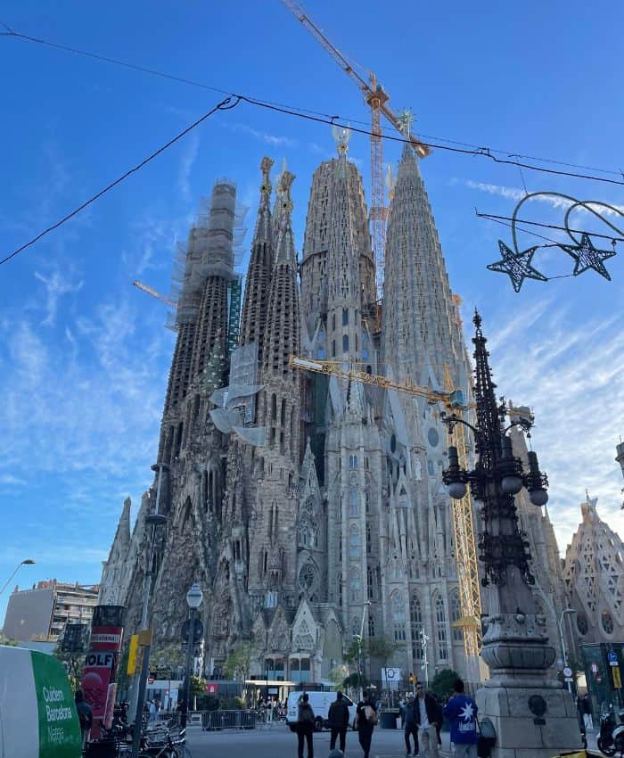 The intricate spires of the Sagrada Familia in Barcelona under construction, with cranes against a clear blue sky, pedestrians, and street decorations in view.