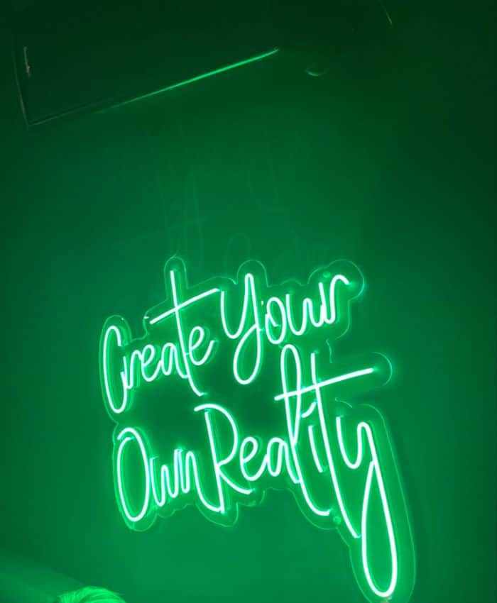 Neon green sign saying 'Create Your Own Reality' on a dark background, with a soft glow around the cursive lettering.