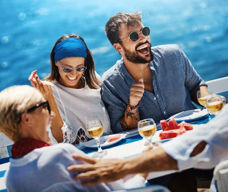 three people laughing at a table on a boat over food and drinks