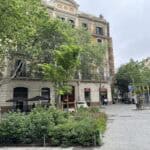 Urban scene with leafy trees, outdoor café seating, and a pedestrian pathway in Barcelona, with a partially visible restaurant sign.