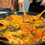 Participants stir a large pan of paella with wooden spoons, mixing vibrant ingredients like rice, red bell peppers, and various vegetables. The hands of several people are visible as they work together.